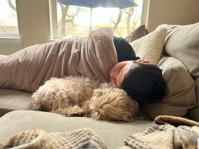 A person sleeping on a couch with a fluffy dog snuggled beside them, both covered by a blanket