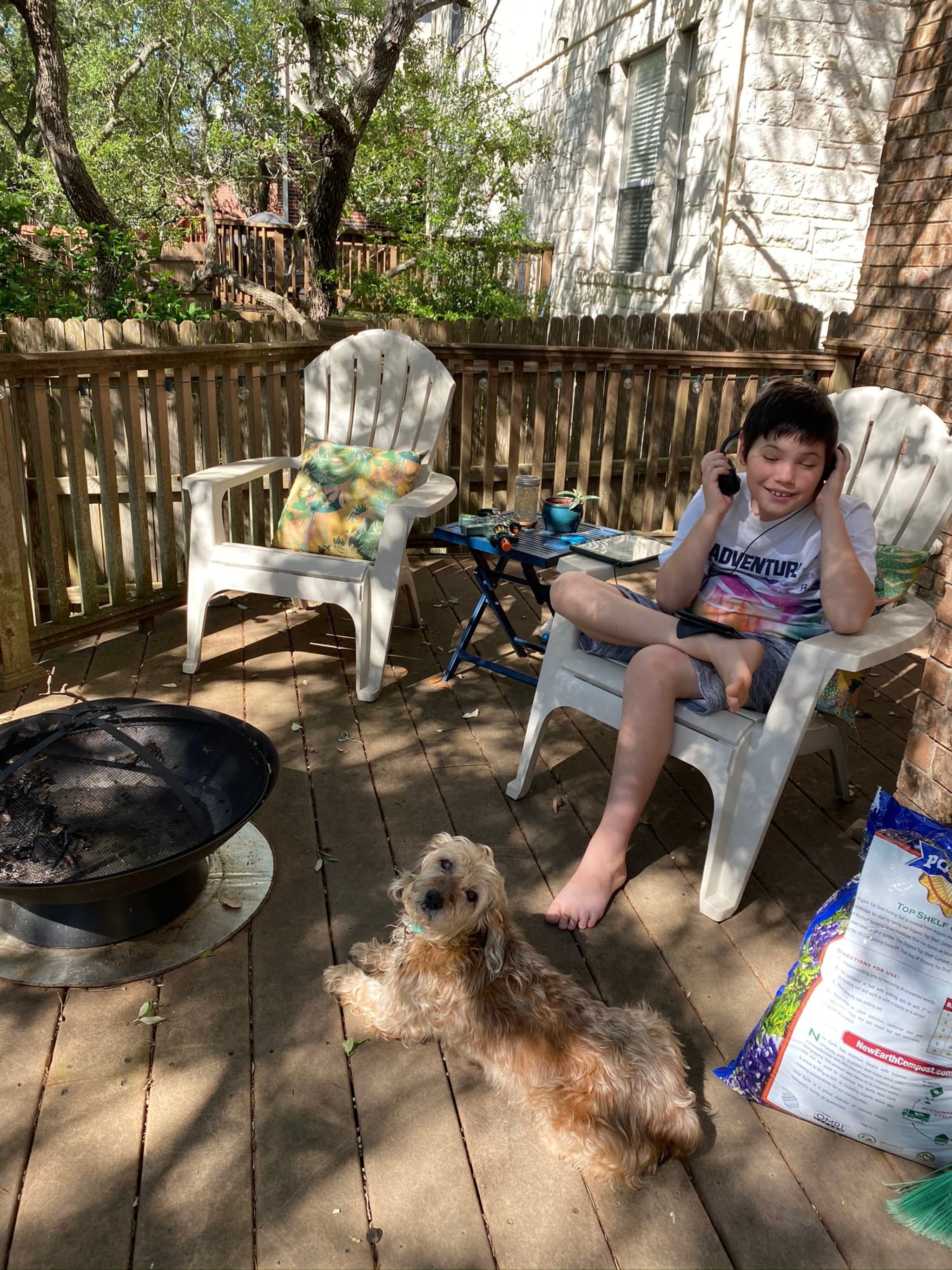 A boy sitting on a chair on a wooden deck, talking on a phone, with a small dog lying nearby. There are two white chairs, a fire pit, and a bag of dog food on the deck, surrounded by trees and a wooden fence
