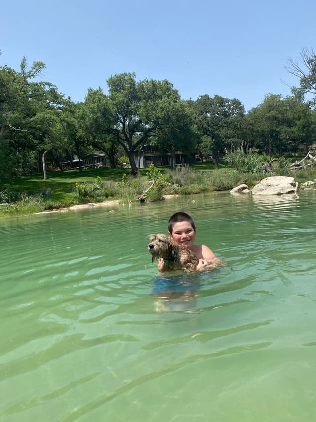 A person holding a small dog while standing in a body of water, with trees and rocks in the background