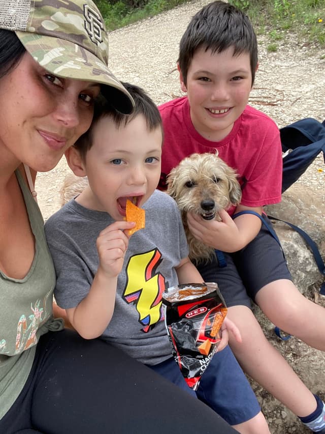A woman and two children sit outdoors on a rock, with one child holding a small dog and the other eating a snack. The woman wears a camouflage cap, and the children are dressed casually