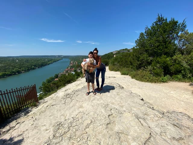 Two people standing on a rocky overlook with a scenic view of a river and greenery under a clear blue sky