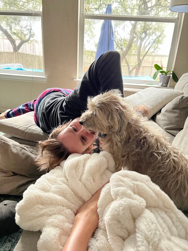 A person lying on a couch with a small dog nearby, surrounded by blankets, in a room with large windows