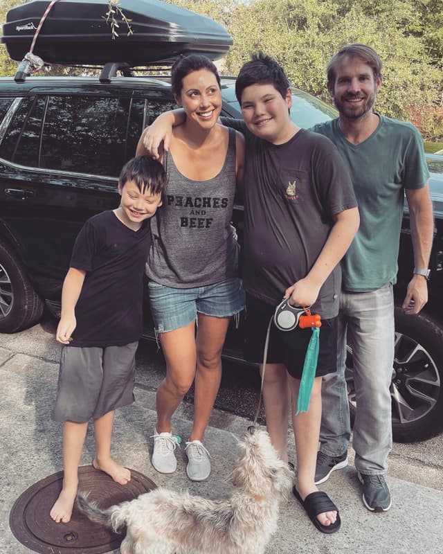 A family of four stands together in front of a car, with a small dog at their feet. The group includes two adults and two children, all casually dressed and smiling