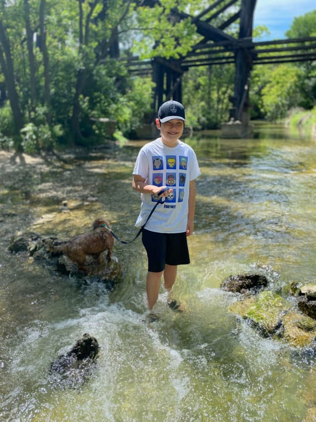 A child wearing a cap and t-shirt stands in a shallow stream, holding a leash attached to a small dog. Trees and a wooden bridge are visible in the background