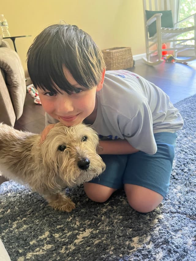 A young boy is kneeling on a carpet, hugging a small, fluffy dog. The room has a cozy atmosphere with a chair and basket in the background