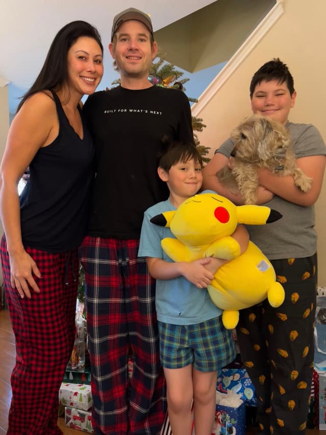 A family of four, dressed in casual clothes, stands together indoors. The youngest child holds a large Pikachu plush toy, and the older child holds a small dog. They are surrounded by wrapped gifts
