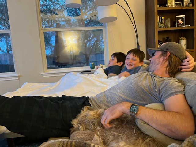 A relaxed scene with two children and an adult lounging on a couch, covered with a blanket, and a dog resting nearby. The room has large windows and a bookshelf in the background
