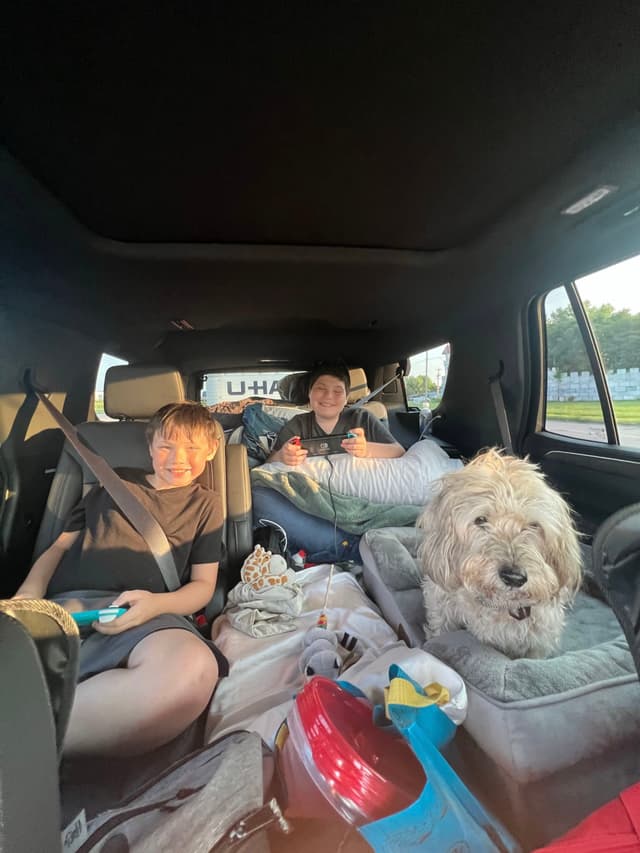 Two children and a dog are in the backseat of a car, surrounded by travel items. The children are smiling, and the dog is sitting on a cushion