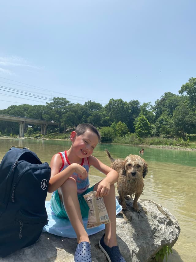 A child and a dog sit on a rock by a river, with a backpack nearby. The child is smiling and holding a snack, while the dog stands beside them. Trees and a bridge are visible in the background under a clear sky