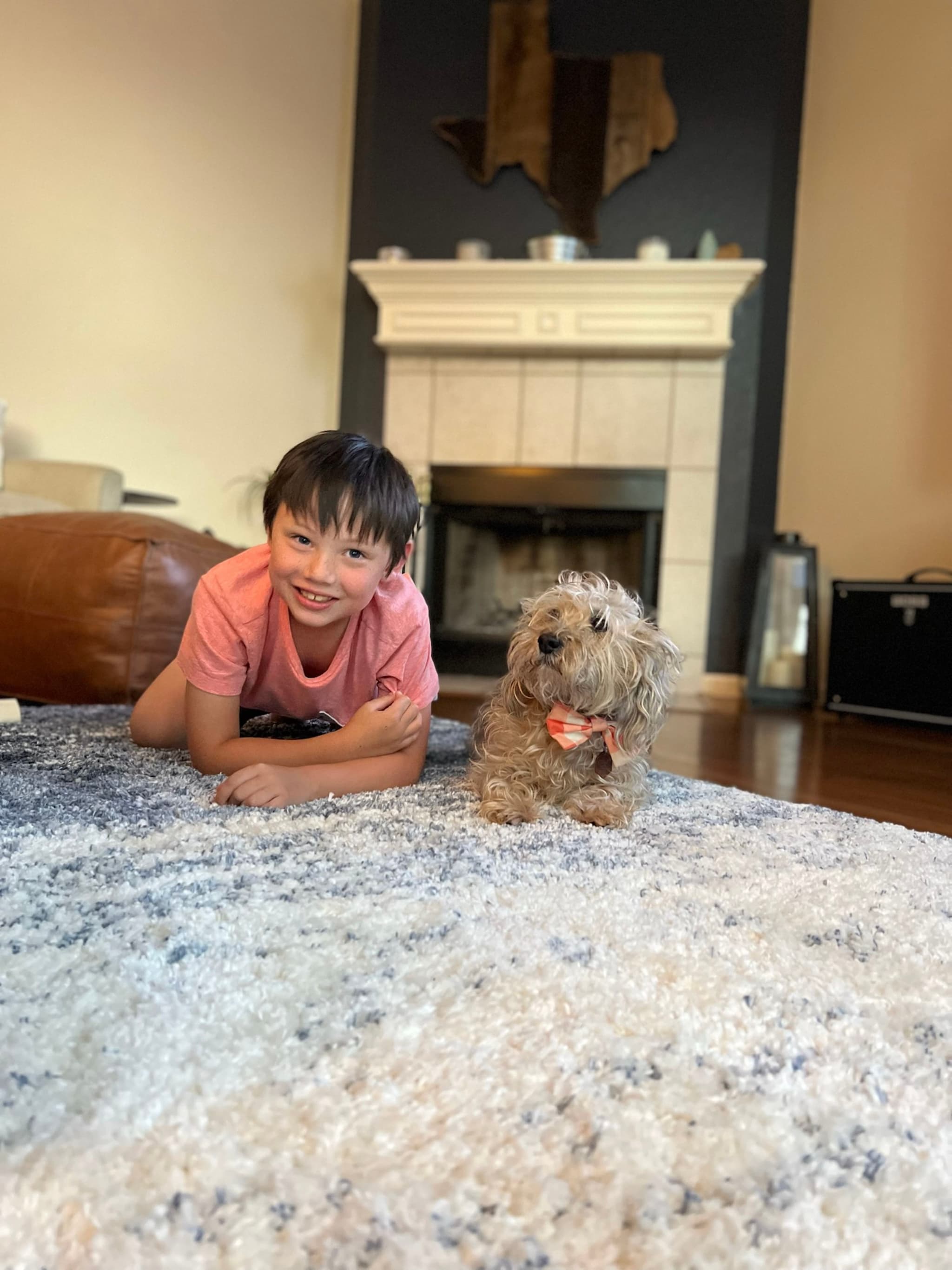 A child in a pink shirt lies on a carpet next to a small dog in a living room with a fireplace in the background