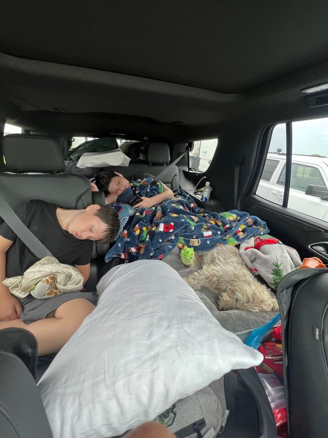 Two children are sleeping in the backseat of a car, surrounded by pillows, blankets, and a small dog