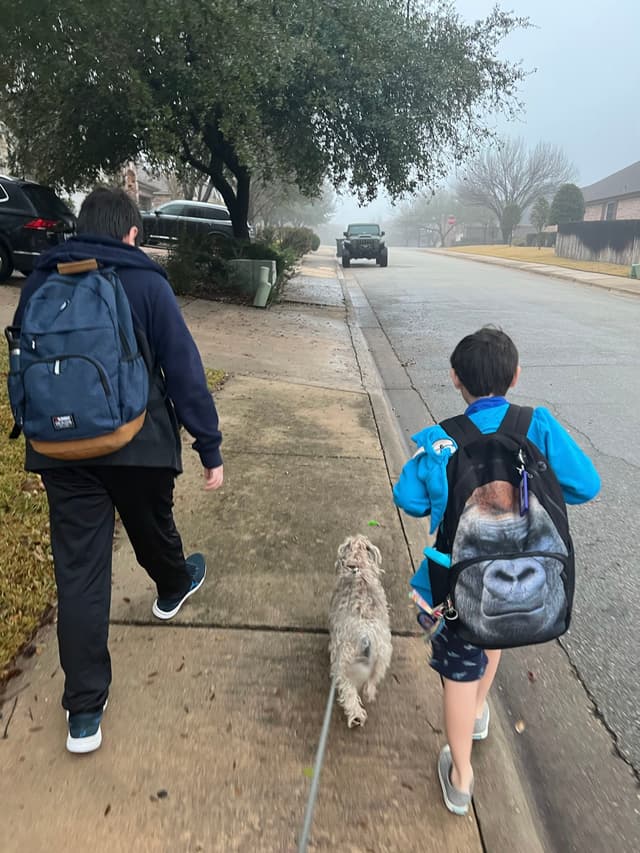 Two people walking a dog on a sidewalk in a suburban neighborhood, both carrying backpacks