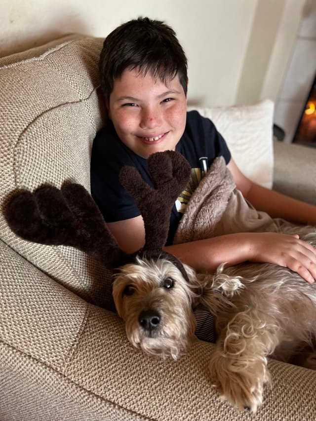 A smiling child is sitting on a couch with a dog wearing reindeer antlers resting beside them