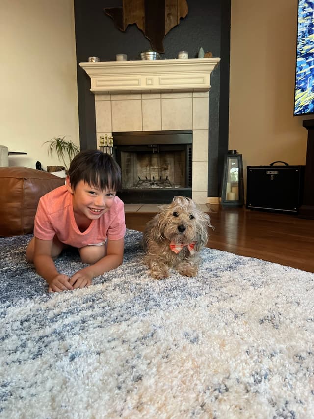 A child in a pink shirt and a small dog with a bow lie on a rug in a living room with a fireplace in the background