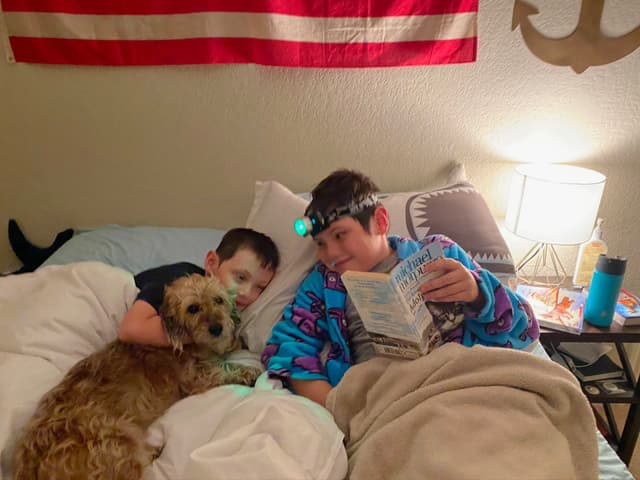 Two children are in bed with a dog, one child is reading a book with a headlamp, and an American flag is hanging on the wall behind them
