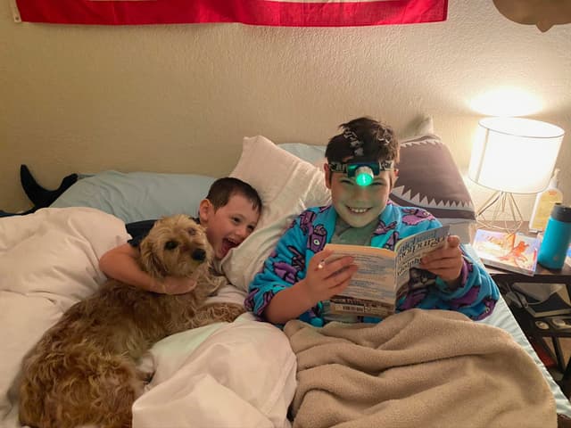 Two children are in bed with a dog. One child is reading a book with a headlamp, while the other is cuddling the dog. A lamp and some items are on the bedside table