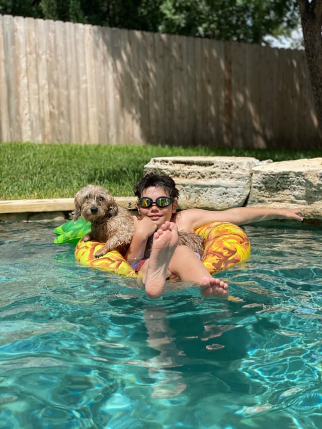 A person wearing sunglasses relaxes on a float in a pool, accompanied by a small dog