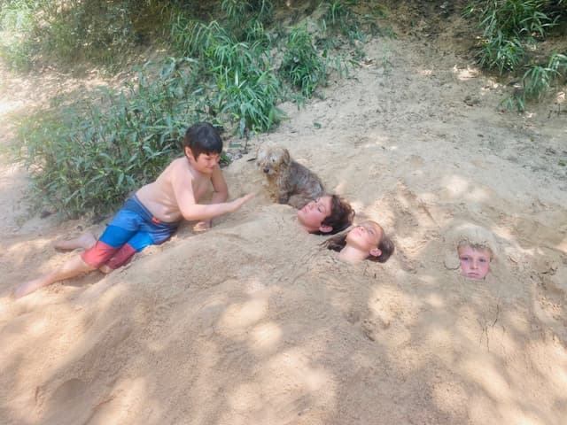 A child lying on sand next to three people buried up to their necks, with a dog sitting nearby