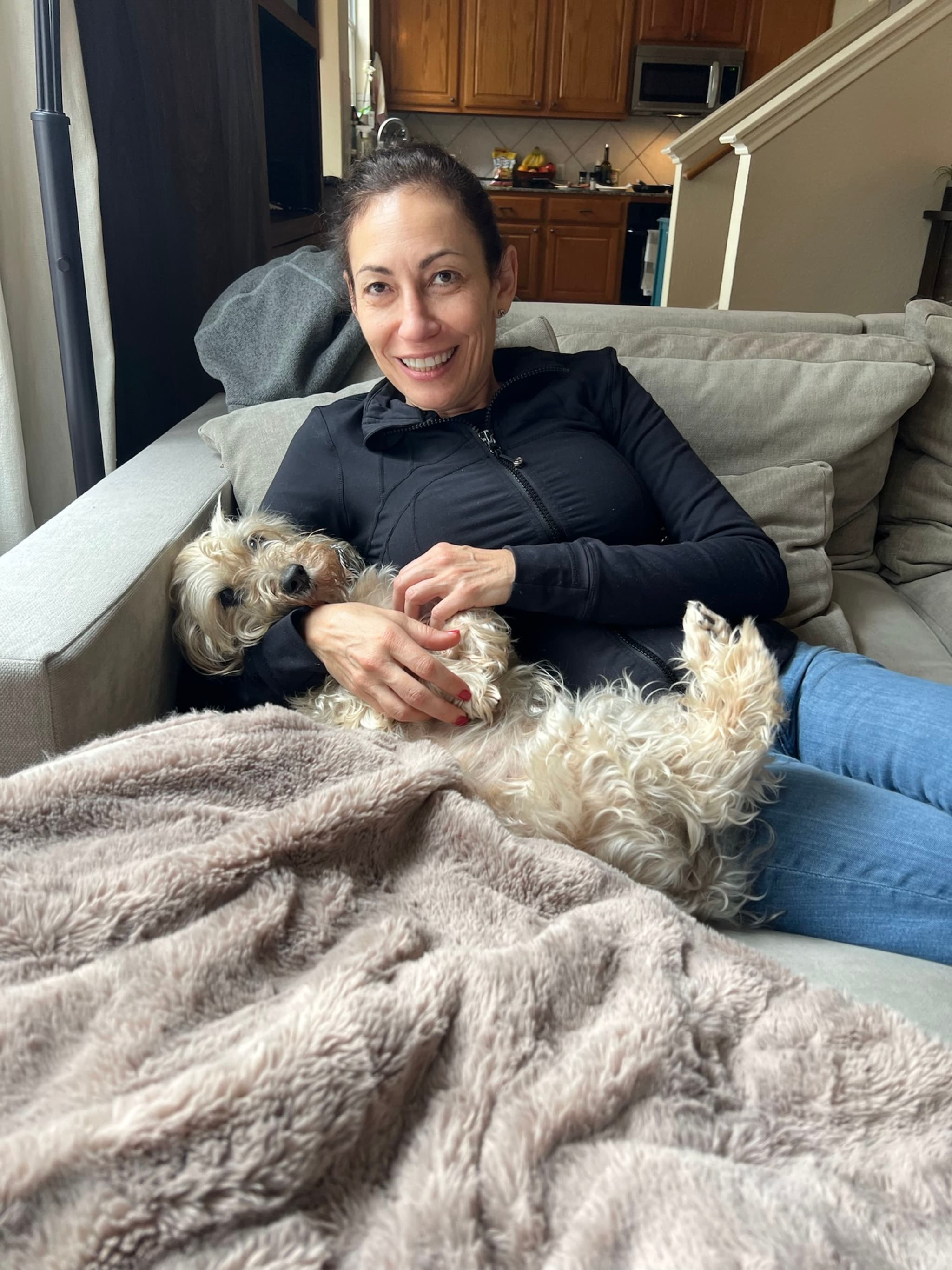A woman is relaxing on a couch with a small dog lying on her lap, covered partially by a fluffy blanket. The background features a kitchen area