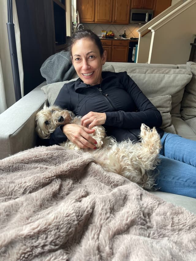 A woman is relaxing on a couch with a small dog lying on her lap, covered partially by a fluffy blanket. The background features a kitchen area