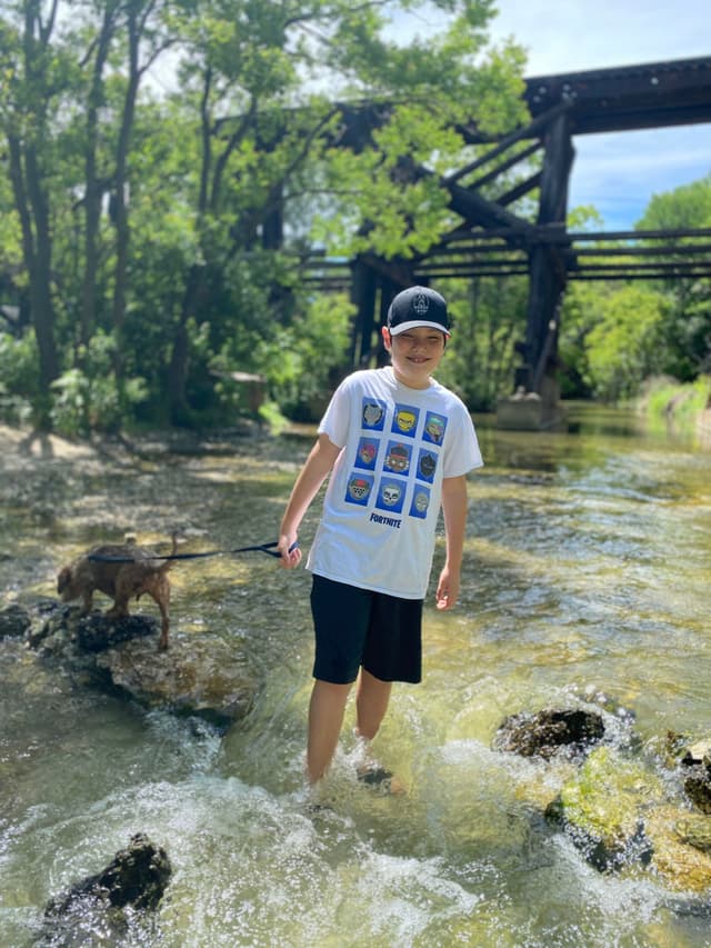 A boy wearing a white T-shirt and black shorts stands in a shallow stream, with a dog nearby on a rock. Trees and a wooden bridge are in the background