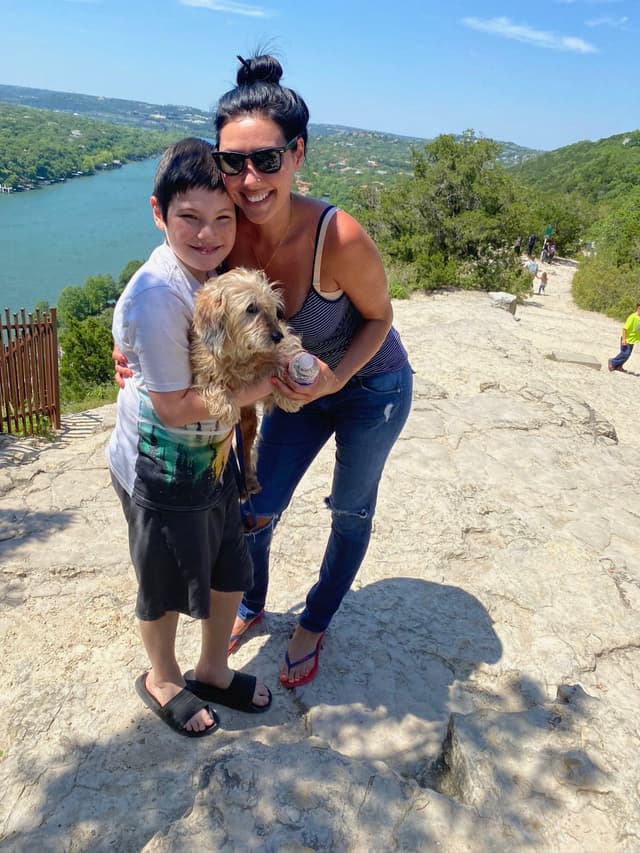A woman and a child are smiling and holding a small dog on a rocky path with a scenic view of a river and greenery in the background