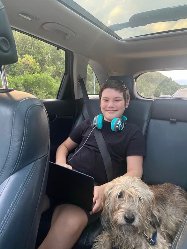 A smiling child with headphones around their neck sits in the backseat of a car, holding a tablet, with a dog resting beside them