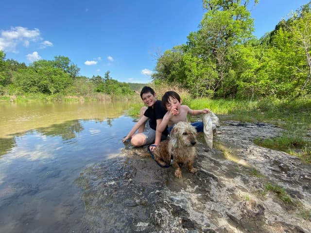 Two people and a dog sitting by a calm river, surrounded by lush greenery and a clear blue sky