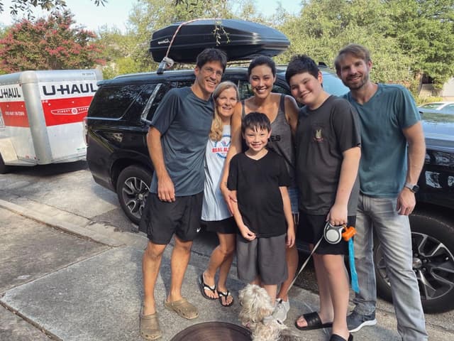 A group of six people, including two children, standing together in front of a black SUV with a U-Haul trailer attached. A small dog is in the foreground