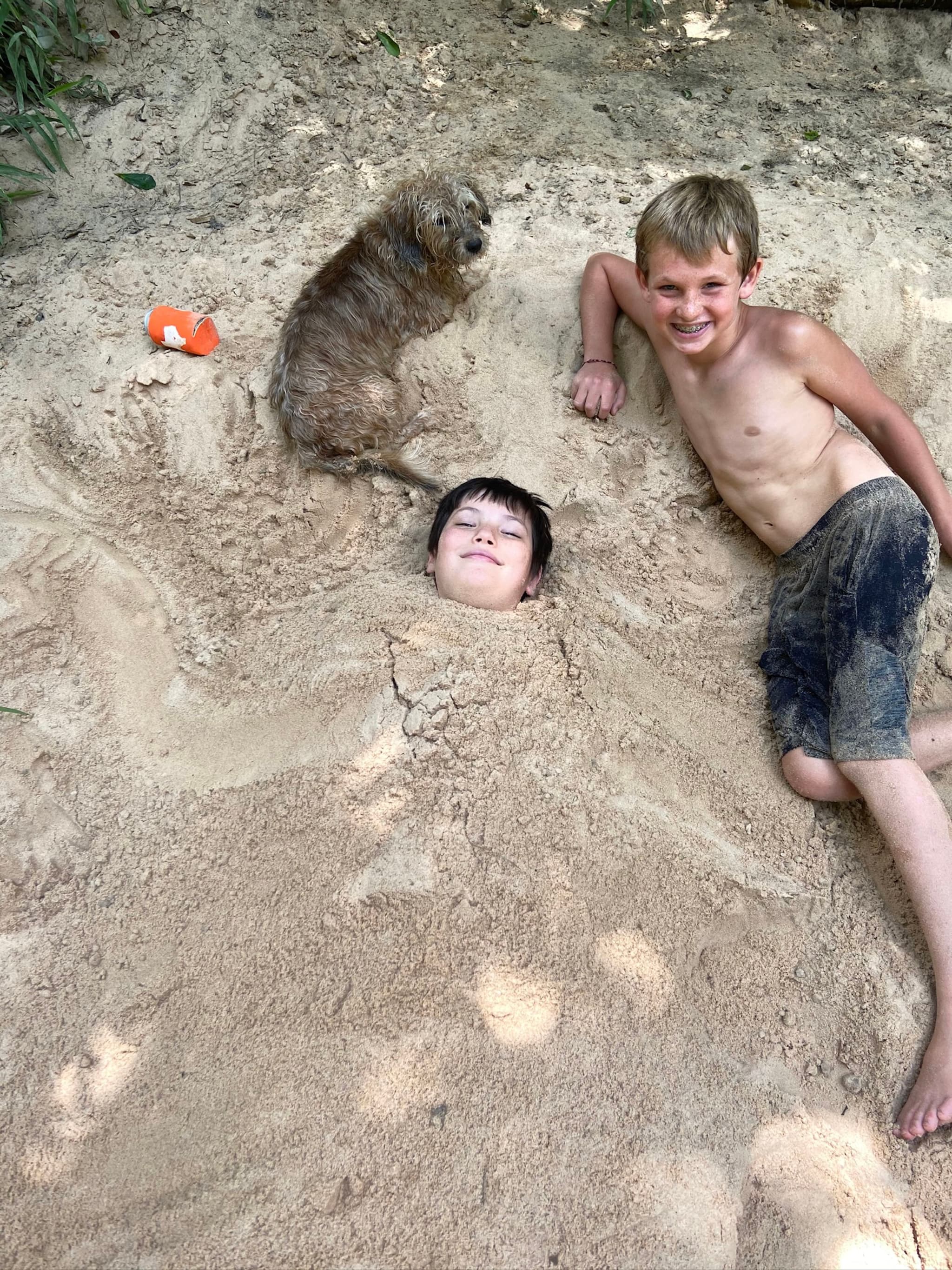 A shirtless boy smiles next to another child buried in sand up to the neck, with a small dog sitting nearby