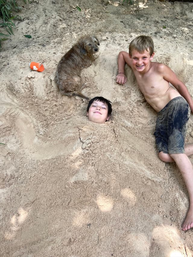 A shirtless boy smiles next to another child buried in sand up to the neck, with a small dog sitting nearby
