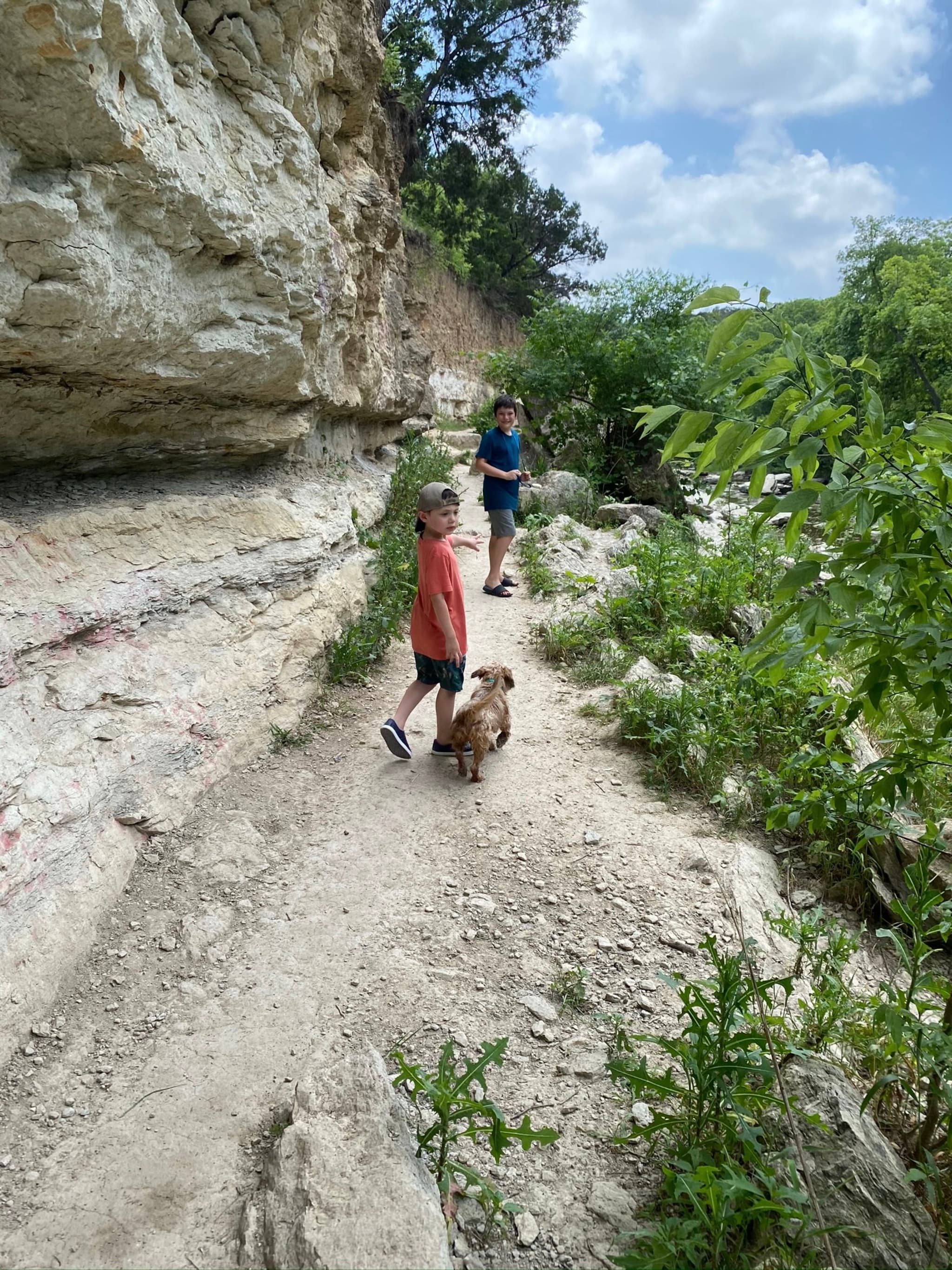 Two children and a dog walking on a rocky trail surrounded by greenery and cliffs under a partly cloudy sky