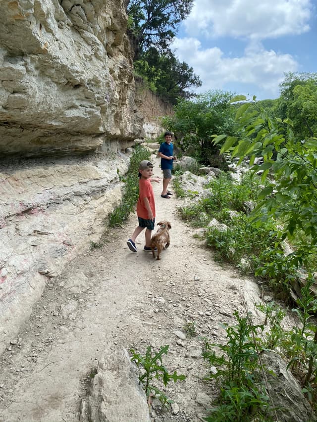 Two children and a dog walking on a rocky trail surrounded by greenery and cliffs under a partly cloudy sky