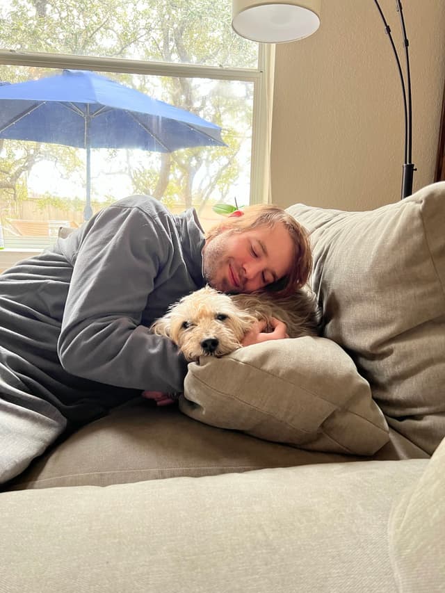 A person is lying on a couch, cuddling with a small dog. The person appears relaxed, with eyes closed, and the dog is resting its head on a pillow. A window in the background shows trees and a blue umbrella outside