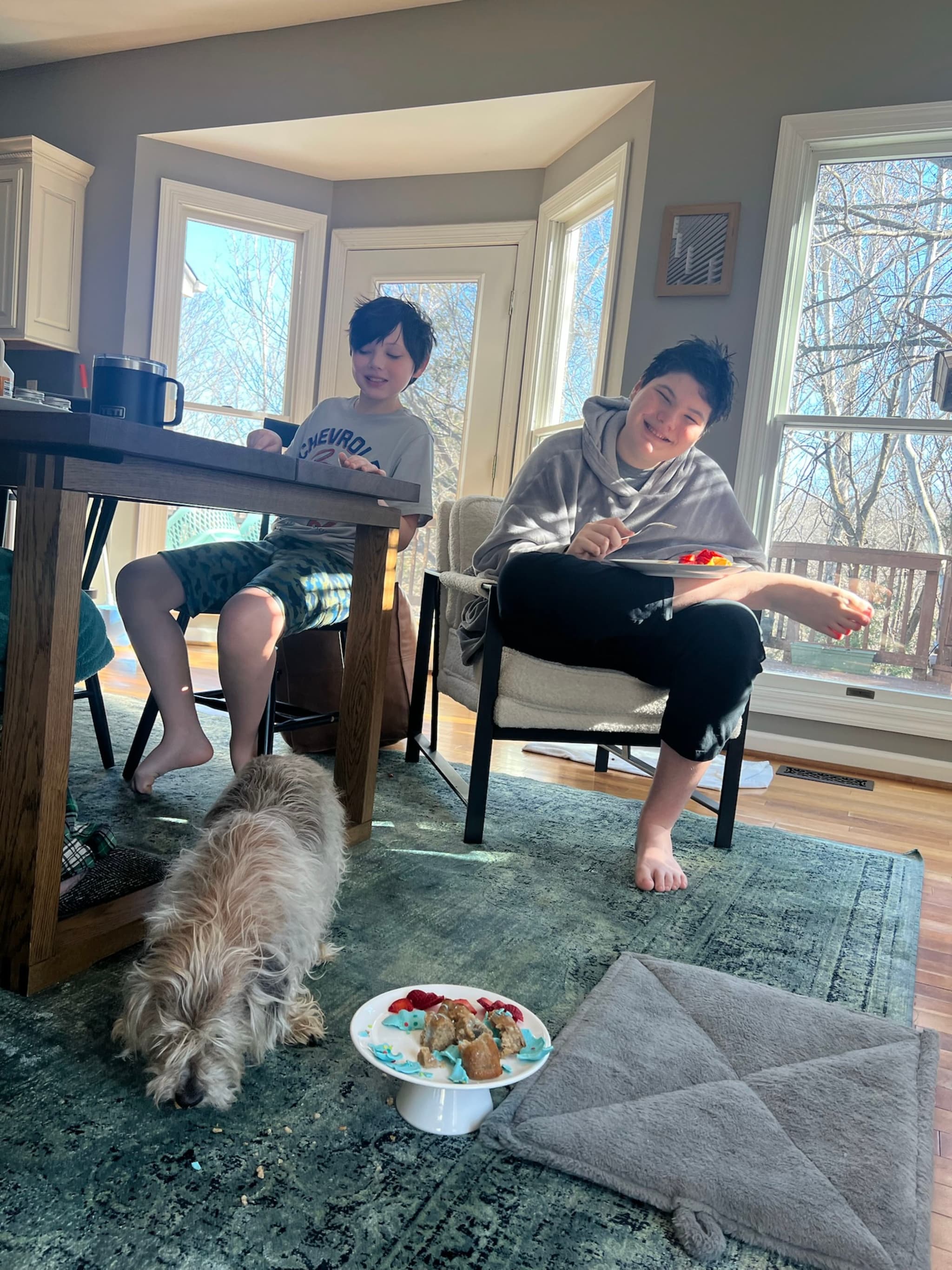 Two people are sitting in a sunlit room, one at a table and the other in a chair, both engaged with their phones. A small dog is on the floor near a bowl of food