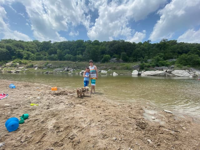 A person and a child stand on a sandy riverbank with toys scattered around. The river flows gently, bordered by lush green trees under a partly cloudy sky