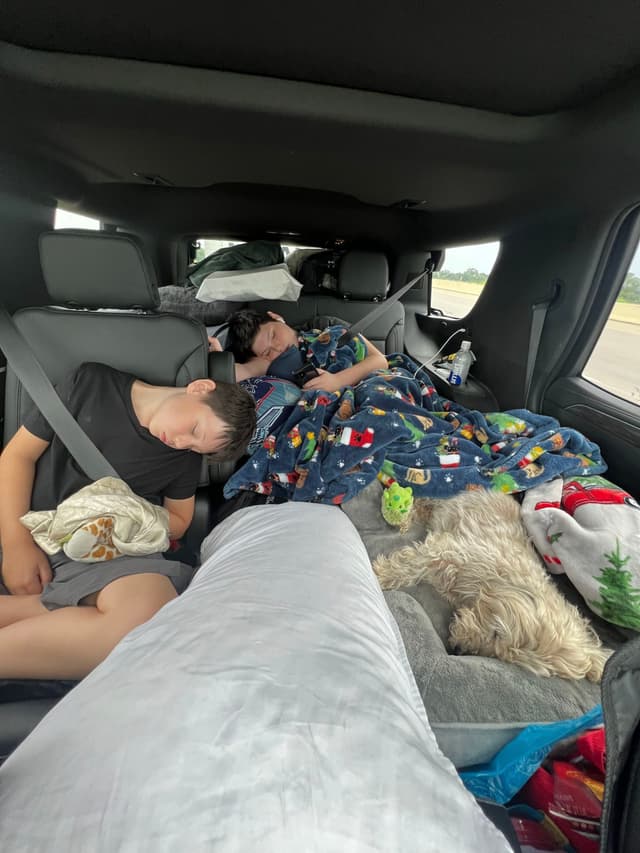 Two children and a dog sleeping in the backseat of a car, surrounded by blankets and pillows