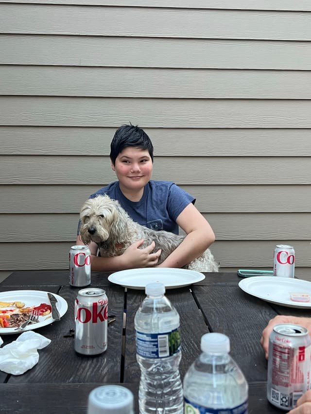 A person sitting at a table with a dog on their lap, surrounded by plates, cans of Diet Coke, and water bottles