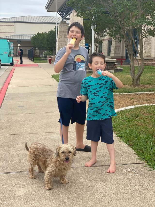 Two children enjoying ice cream stand on a sidewalk with a small dog in front of them