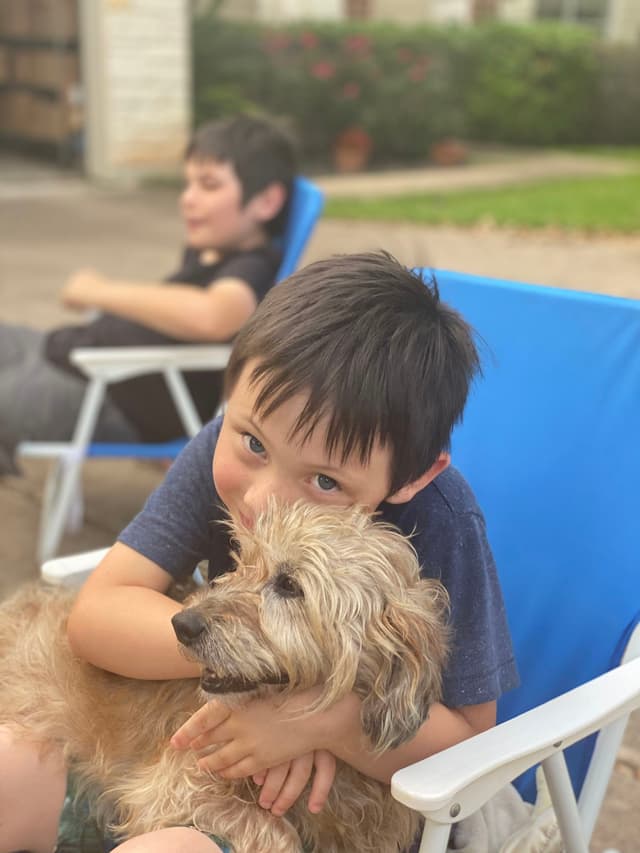 A child sitting on a blue chair hugs a fluffy dog, with another child relaxing in the background on a similar chair
