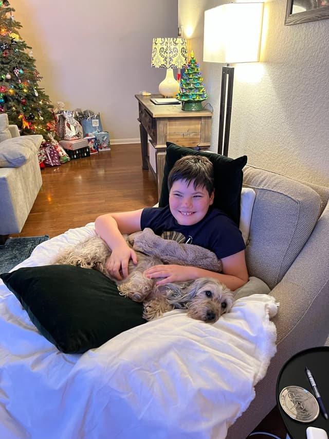 A child is sitting on a cozy chair, smiling and holding a small dog. The room is decorated with a Christmas tree and festive ornaments. A lamp and a small table with a tree-shaped decoration are in the background