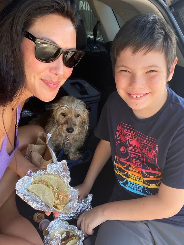 A woman and a boy are sitting in a car trunk, smiling and holding food wrapped in foil. A small dog is sitting between them