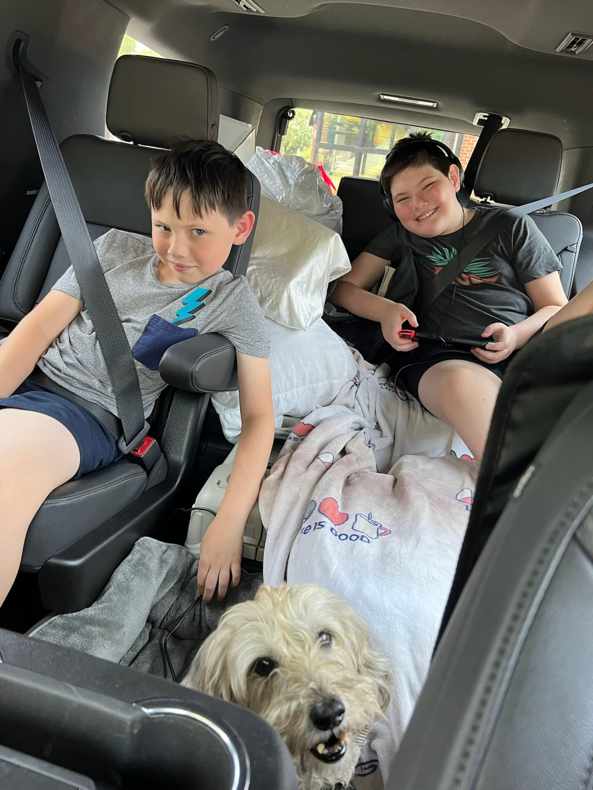 Two children are sitting in the backseat of a car, surrounded by pillows and blankets, with a dog sitting between them