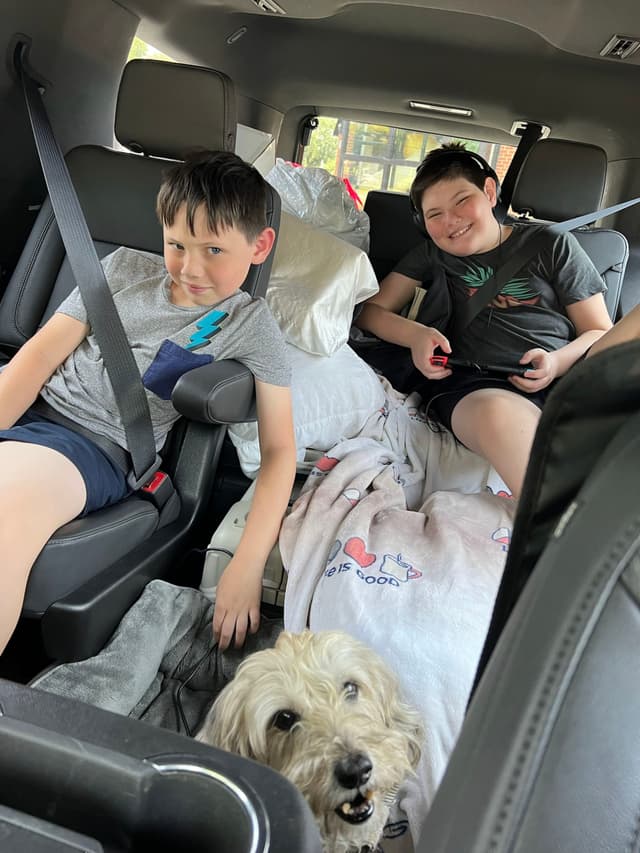 Two children are sitting in the backseat of a car, surrounded by pillows and blankets, with a dog sitting between them