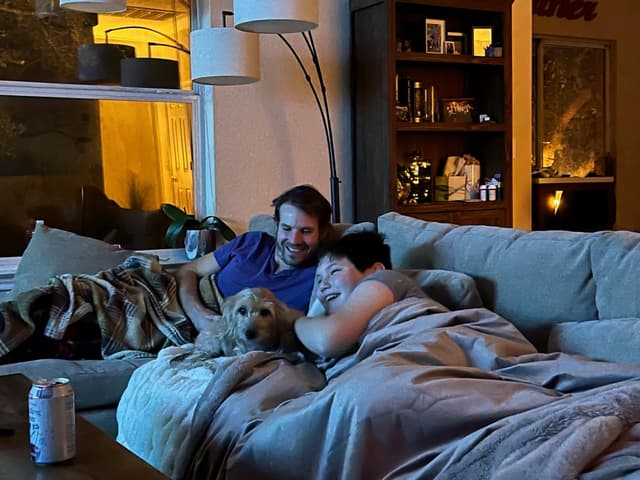 A cozy scene with two people and a dog snuggled under blankets on a couch, illuminated by warm indoor lighting