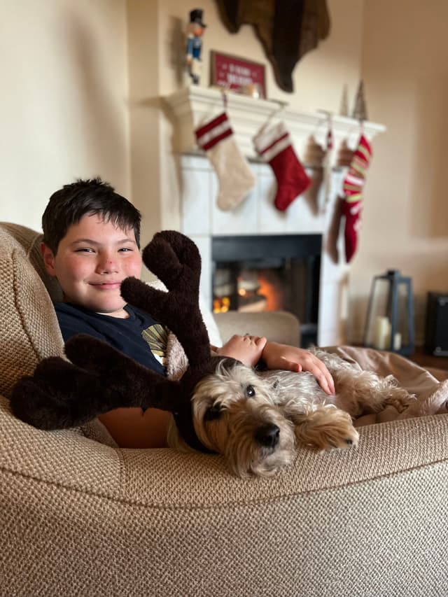 A boy sitting on a couch with a dog wearing reindeer antlers, in front of a fireplace with Christmas stockings hanging above