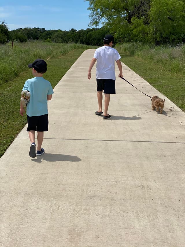 Two people walking on a paved path, one holding a small dog on a leash, surrounded by grass and trees