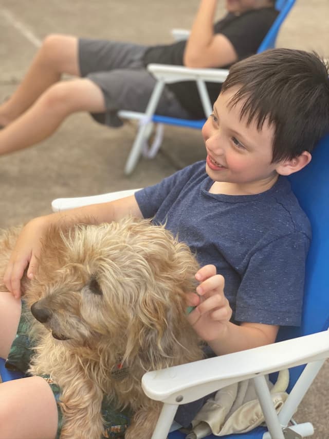 A young boy sitting in a blue chair, smiling and petting a fluffy dog on his lap, with another person sitting in the background