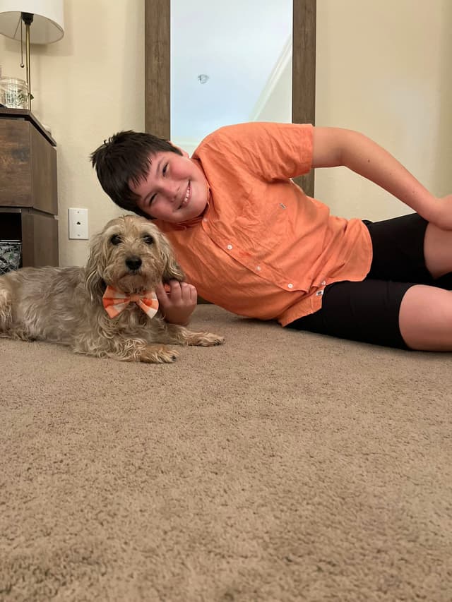 A boy in an orange shirt is lying on the carpet next to a small dog wearing a matching orange bow tie