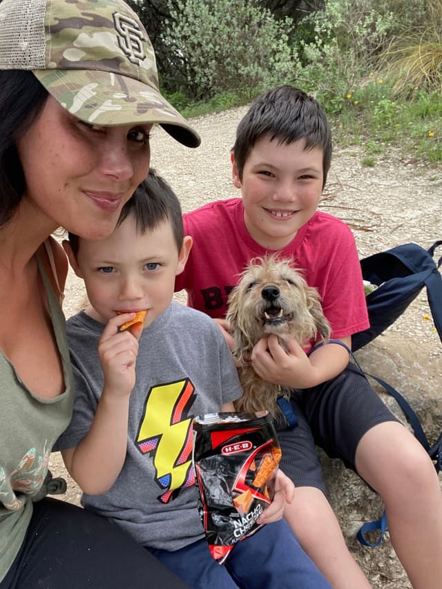 A woman and two children are sitting outdoors on a trail, with one child holding a small dog. The younger child is eating from a bag of chips. The woman is wearing a camouflage cap, and they all appear to be enjoying a casual moment in nature